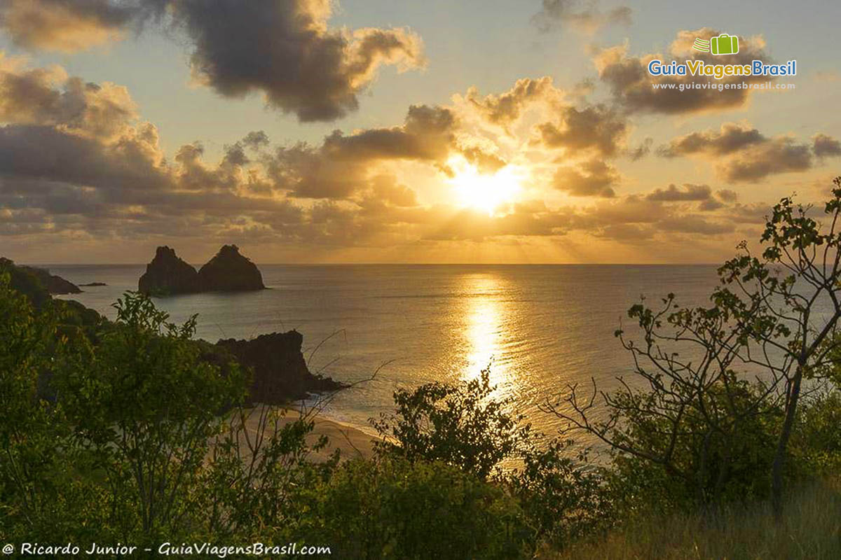 Imagem do sol de pondo no meio e do lado esquerdo o Morro Dois Irmãos, na Praia do Boldro, em Fernando de Noronha, Pernambuco, Brasil.